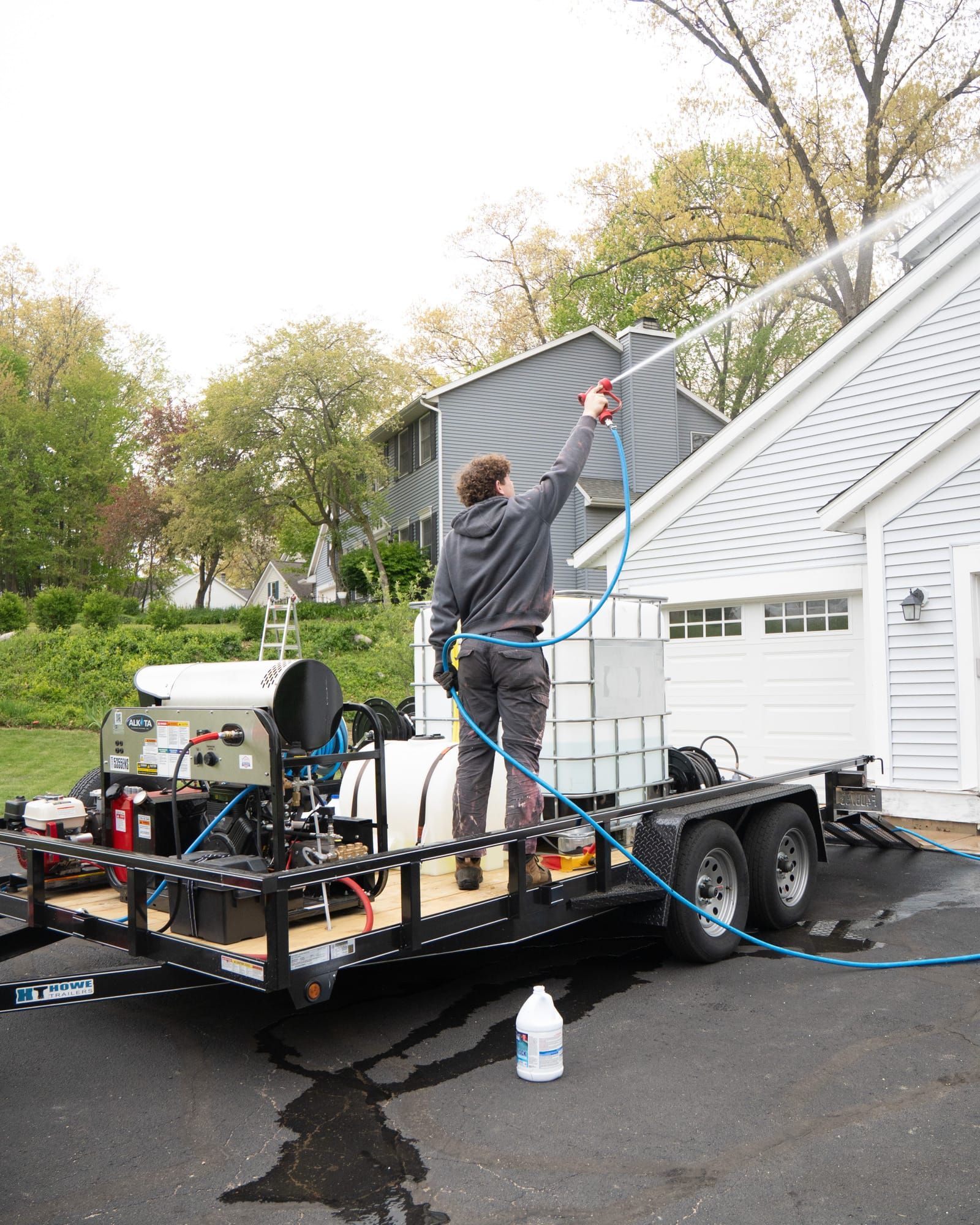 Elevation technician soft-washing a roofline from the trailer rig, with pressure washer, water tank and hose reel aboard